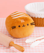 Front view of Pusheen Loaf Squisheen. The light brown plush is in the shape of a 3D oval. The front of the plush features brown embroidery details with light brown cat ears extending off the top. On the top of the squisheen are cream-colored wavy lines to mimic bake lines. The plush sits on a pink and white surface surrounded by a breadbasket, rolling pin, and cream tongs.  