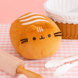 Front view of Pusheen Loaf Squisheen. The light brown plush is in the shape of a 3D oval. The front of the plush features brown embroidery details with light brown cat ears extending off the top. On the top of the squisheen are cream-colored wavy lines to mimic bake lines. The plush sits on a pink and white surface surrounded by a breadbasket, rolling pin, and cream tongs.  