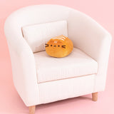 Pusheen Loaf Squisheen sits on a cream chair to show the size of the plush. The light brown bun loaf-shaped plush has brown embroidery features for Pusheen's eyes, mouth, and whiskers.