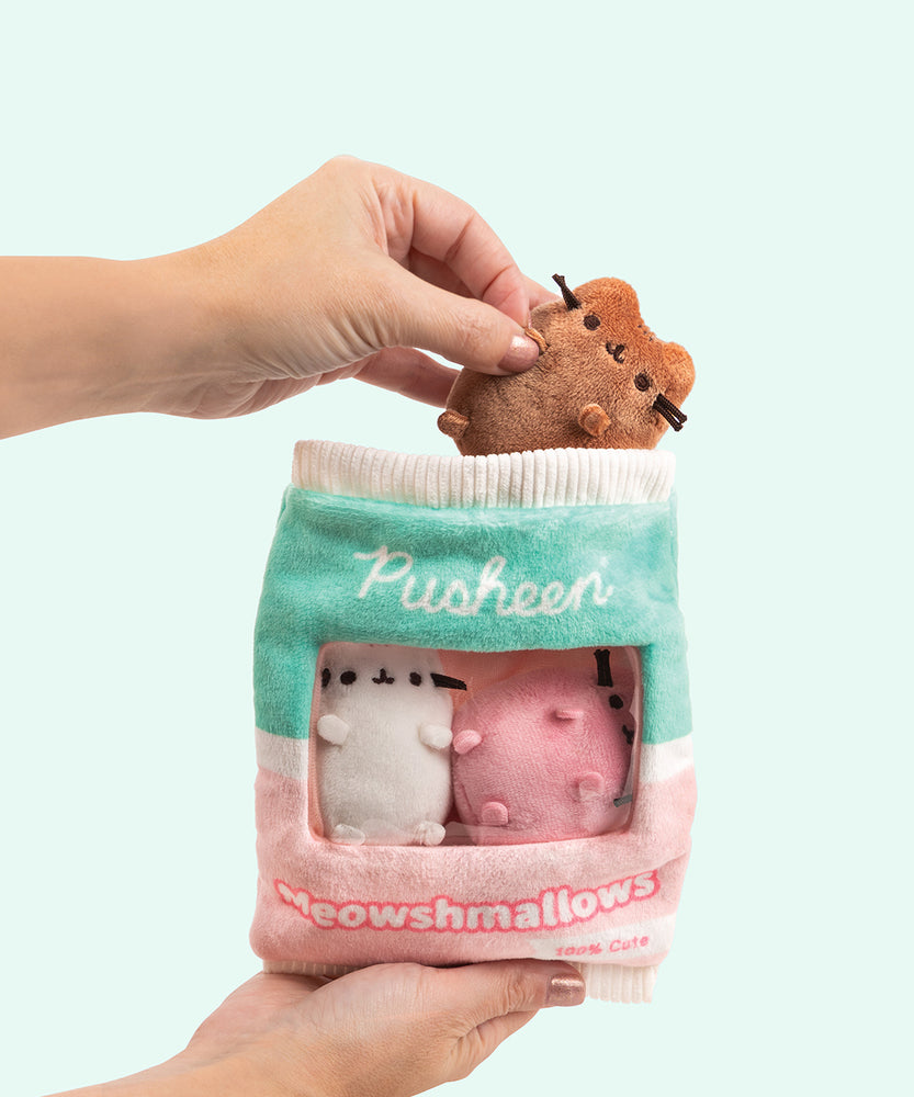 Hand holds the brown Pusheen Meowshmallow out of the plush bag containing white and pink Meowshmallows. Another hand holds the Pusheen Meowshmallows bag has a white seal, mint green top portion, and light pink bottom portion. 