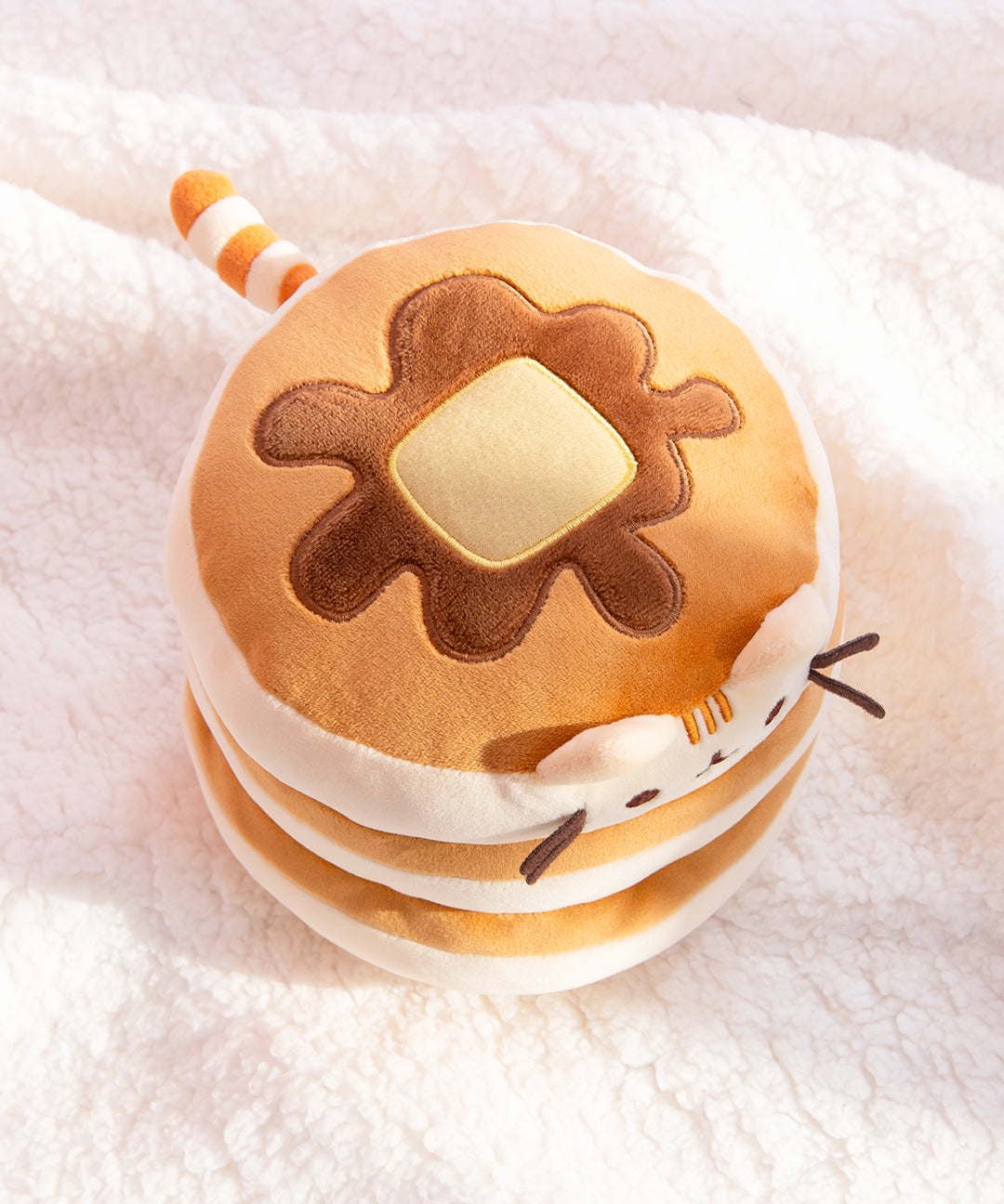 Top view of the Pusheen Pancake Squisheen on a white fluffy background. The pad of butter and syrup drizzle edged with light yellow and dark brown embroidery respectively. Pusheen's cream and light brown striped tail extends from the back of the stacked plush. 