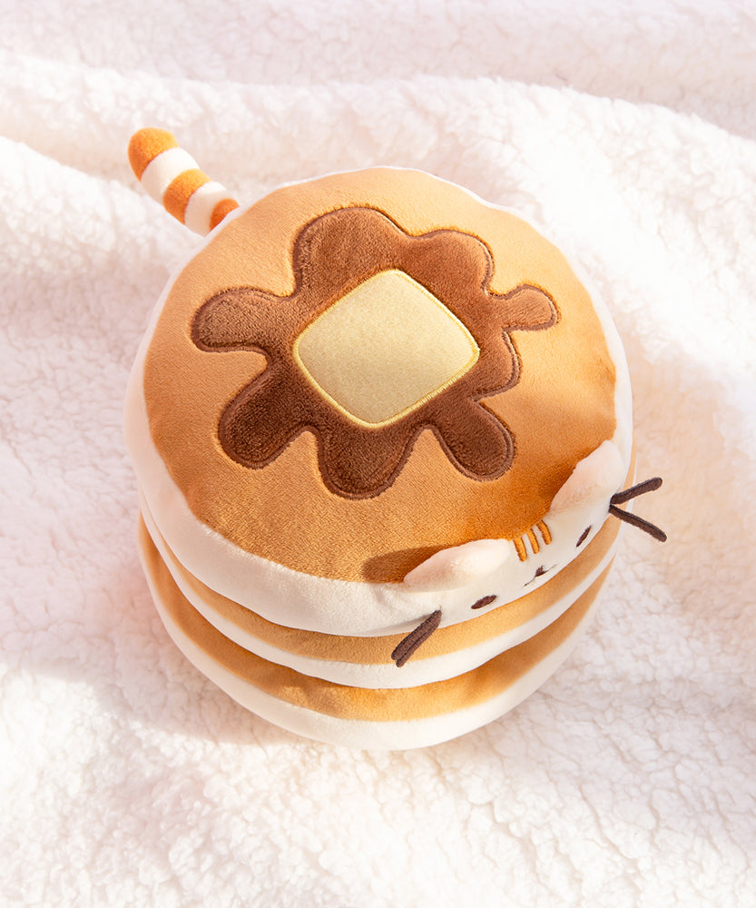 Top view of the Pusheen Pancake Squisheen on a white fluffy background. The pad of butter and syrup drizzle edged with light yellow and dark brown embroidery respectively. Pusheen's cream and light brown striped tail extends from the back of the stacked plush. 