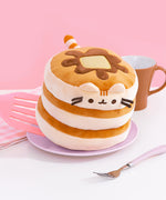 Front view of a cylinder plush that resembles a pancake stack with a pad of butter and syrup drizzle. Pusheen's ears are light brown and sit near the top of the stack and the three light brown embroidered stripes are between her ears. The plush resembles a stack of pancakes and is alternating cream and light brown coloring.