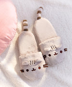 Pusheen Plush Slippers lying on a white fluffy surface next to a light pink pillow. Plush slippers have Pusheen's face embroidered accompanied by ears that extend off the toe box. Off the back of the slippers are Pusheen's striped tails. 