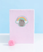 Front view of Pusheen Rainbow Plush Notebook. The pink notebook is made of a fuzzy pink material. The notebook includes a pink pom pom bookmark. On the front center of the notebook is an emblem of Pusheen the Cat licking her paw while surrounded by a pink, yellow, and blue rainbow with white clouds.  