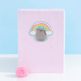 Front view of Pusheen Rainbow Plush Notebook. The pink notebook is made of a fuzzy pink material. The notebook includes a pink pom pom bookmark. On the front center of the notebook is an emblem of Pusheen the Cat licking her paw while surrounded by a pink, yellow, and blue rainbow with white clouds.  