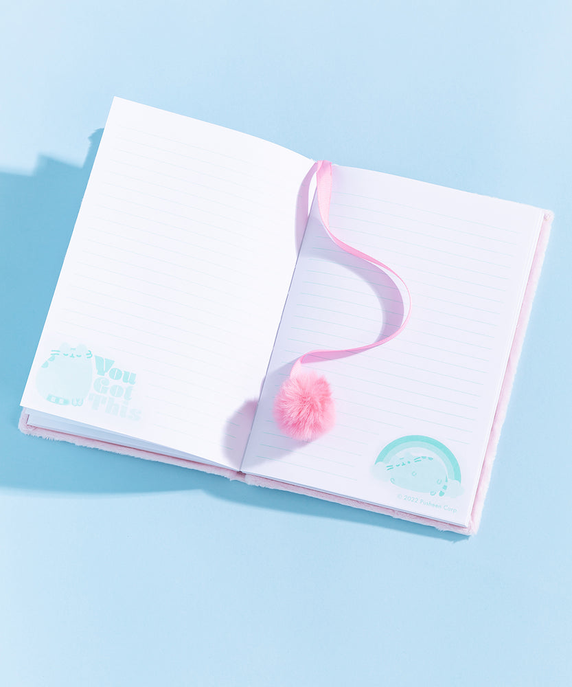 The Pusheen Rainbow Plush Notebook lies on a light blue surface. In this view, the texture of the light pink fluffy plush surface can be seen in more detail.  