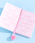 Interior view of plush notebook. The protective first page is shades of pink and features a pattern of Pusheen posed two ways with rainbows and clouds. The pink pom pom bookmark lies in front of the open notebook.  