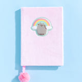 The Pusheen Rainbow Plush Notebook lies on a light blue surface. In this view, the texture of the light pink fluffy plush surface can be seen in more detail.  