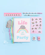 Front view of all the contents that come in the Pusheen Rainbow Stationery set. The clear zip pouch, notebook, sticker sheet, pencil, pencil sharpener, pen, eraser, and ruler sit atop white cubes in front of a light blue background.  