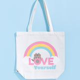 Pusheen Rainbow Tote Bag lies on a light blue surface. The white canvas tote bag features a grey Pusheen the Cat surrounded by a pink, yellow, and blue rainbow and white clouds. Pusheen sits atop the pink and blue colored phrase “Love Yourself.” 
