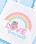 Pusheen Rainbow Tote Bag lies on a light blue surface. The white canvas tote bag features a grey Pusheen the Cat surrounded by a pink, yellow, and blue rainbow and white clouds. Pusheen sits atop the pink and blue colored phrase “Love Yourself.” 