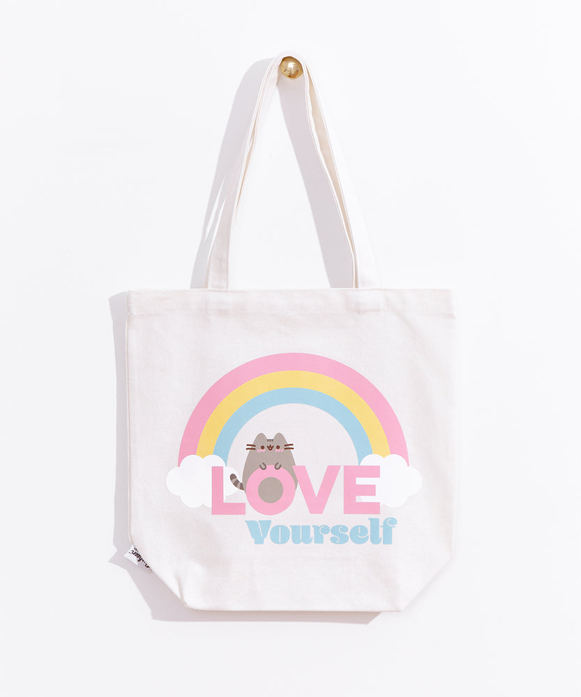 The white canvas bag with pink, yellow, blue, white, grey, and brown printed graphic hangs on a gold knob in front of a white background. 