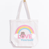 The white canvas bag with pink, yellow, blue, white, grey, and brown printed graphic hangs on a gold knob in front of a white background. 