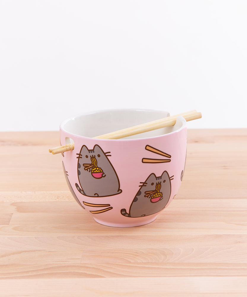 The Pusheen Ramen Bowl with the chopsticks resting in the holes on top of a wooden countertop in front of a white background. The top portion of the chopstick rests on the intent of the bowl’s rim, while the tips of the chopstick slide into the square hole on the opposite side, allowing the chopsticks to rest across the middle of the bowl.  