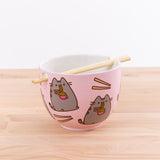 The Pusheen Ramen Bowl with the chopsticks resting in the holes on top of a wooden countertop in front of a white background. The top portion of the chopstick rests on the intent of the bowl’s rim, while the tips of the chopstick slide into the square hole on the opposite side, allowing the chopsticks to rest across the middle of the bowl.  