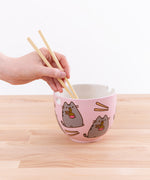 A hand holding a pair of plastic chopsticks into a pink bowl featuring a repeating pattern of chop sticks and Pusheen eating a bowl of ramen. The bowl has two holes around the edges, one square hole on one edge, and a square indent on the rim of the bowl on the opposite side. 
