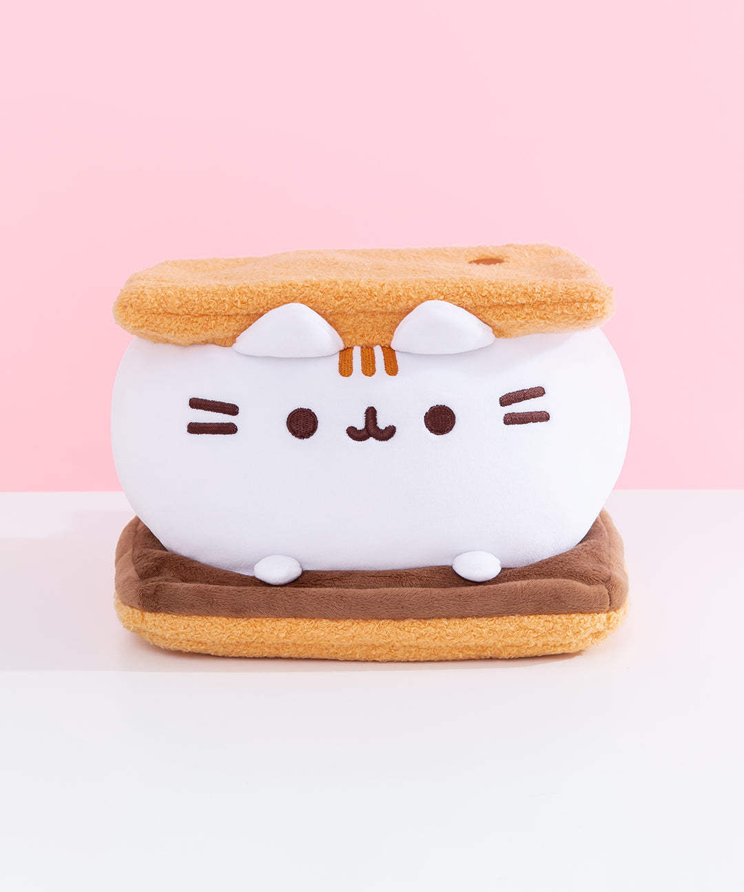 Front view of Pusheen S’mores Squisheen. Pusheen the Cat takes the form of a delicious campfire snack: a s’more. Pusheen's body is the white middle marshmallow. The white marshmallow sits on a dark brown slab of chocolate. The marshmallow is sandwiched between two light brown graham crackers inspired plush.