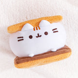 S’mores-inspired plush laying down on a white fluffy backdrop. The Squisheen marshmallow element is white, the graham cracker plush are light brown, and the chocolate slab is a dark brown color.
