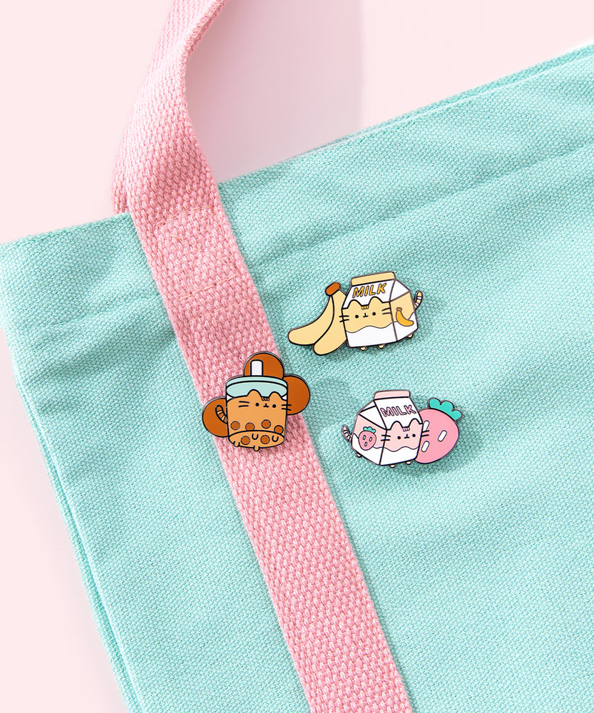 A close-up view of the three pins lying on a mint and pink tote bag. The pins feature milk cartons, a boba tea, bananas, a strawberry, and boba pearls.  Pusheen Sips Pin Set and backer card sits in front of white pedestals and a wire basket. The pin set is surrounded by fuzzy pink, teal, and yellow puffs. The colors in the pin set are pink, white, brown, mint, and yellow 
