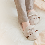 Model wearing the Pusheen Plush Slippers and grey pajama pants nestled on top of a bed, with some apricot sheets on the right hand side.