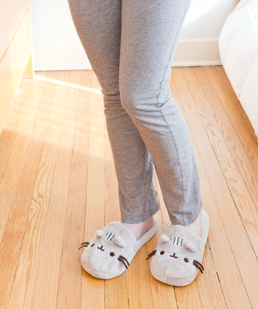 Model wearing the Pusheen Plush Slippers, wearing loose grey pajama pants and standing on top of a wooden floor.