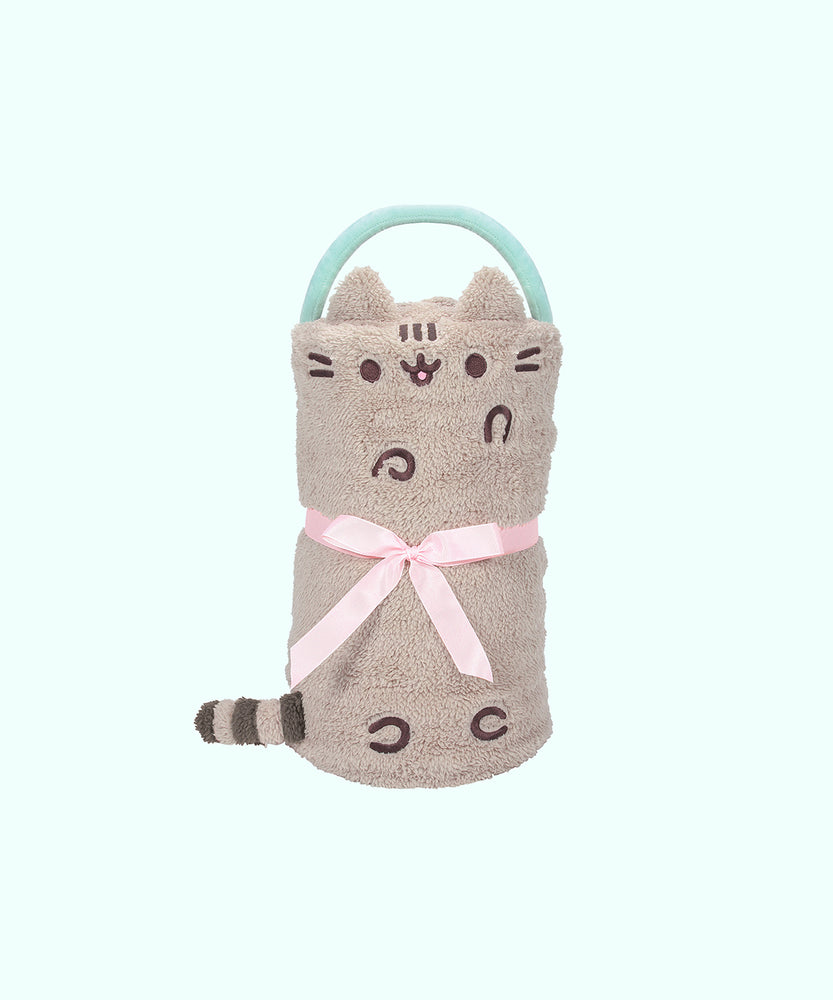 Front view of the Pusheen Snowthrow. This folded blanket is tied with a light pink ribbon to fold the blanket into a spherical shape. The throw blanket includes a mint green handle for easy traveling. On the front of the light grey sherpa features Pusheen the Cat's classic features of paws, whiskers, and her facial features embrodiered in a brown thread. Pusheen's signature striped grey and brown tail extends off the side of the blanket.