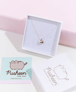 The silver charm necklace in a square white jewelry box, propped up against a pink pedestal, the square card insert and the lid of the box laying underneath it. The square card features an illustration of the Pusheen the Cat logo.