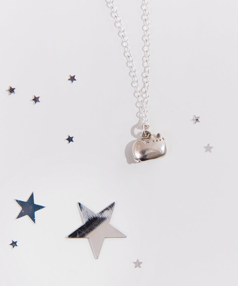 Close up of the silver Pusheen charm necklace, surrounded by reflective silver stars of various size. The details of Pusheen’s face are carved into the charm, while her ears and tail are molded as part of the charm. The loop connecting the charm to the chain necklace is on top of pusheen’s back.