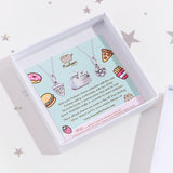 The back of the square card insert, featuring a photo of the silver Pusheen charm, alongside some copy regarding other charms from the manufacturer licensed to charm. Illustrations of various foods decorate the edges of the card.