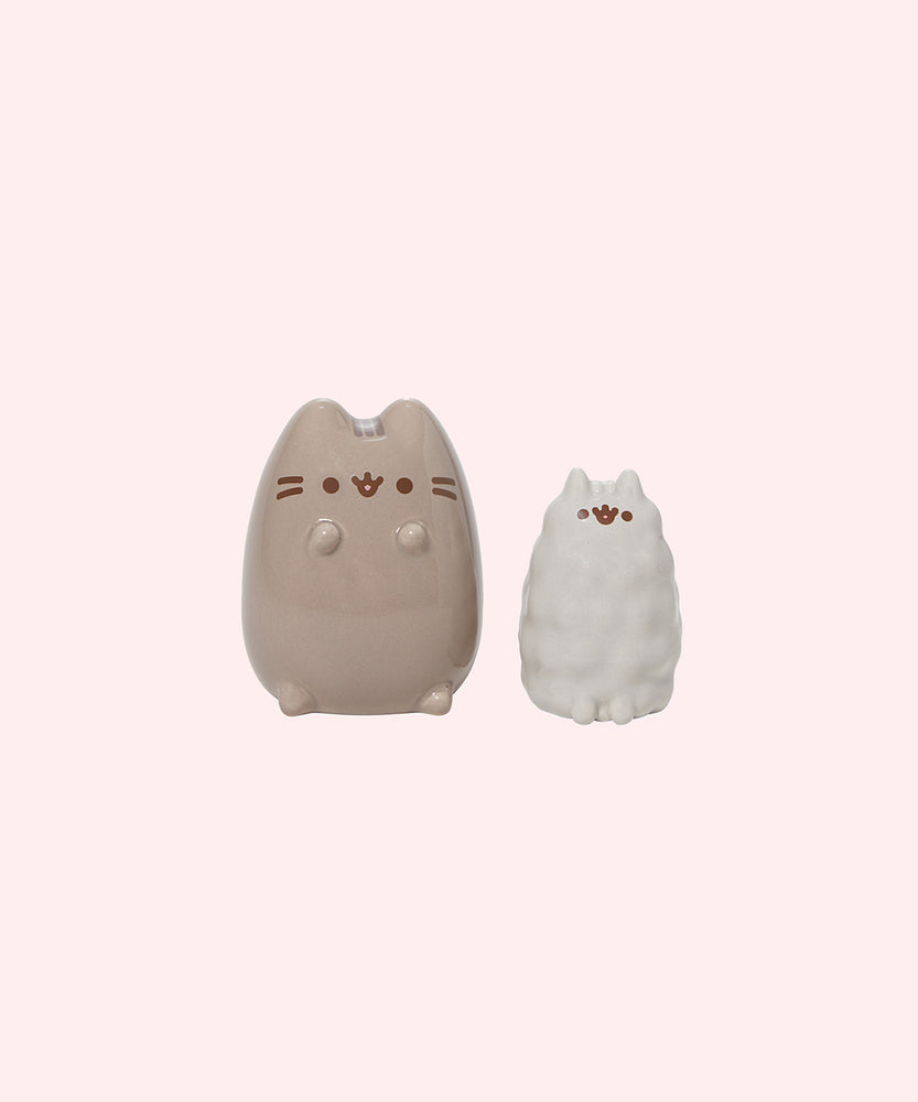 Front view of the Pusheen & Stormy Salt & Pepper Set. These ceramic shakers are in the shape of Pusheen the Cat and little sister Stormy.  Pusheen is shown in a darker grey brown and has her classic facial features and whiskers printed on front with her paws slightly extending off the body to remain seated. The Stormy shaker is seated on the right of Pusheen, and she is light grey with a smooth featured appearance to the ceramic to mimic fur.