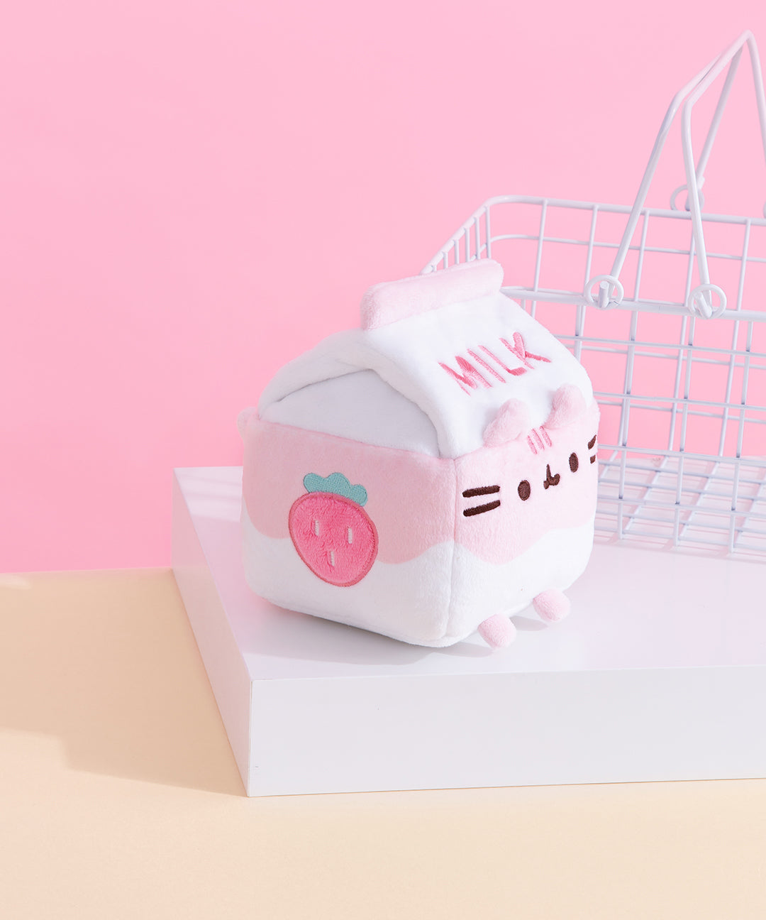 A side profile of the Strawberry Milk Sips Plush shows off the pink strawberry embroidered on the side of the plush. The plush is seated on a white pedestal with a small white wire basket in the background.  