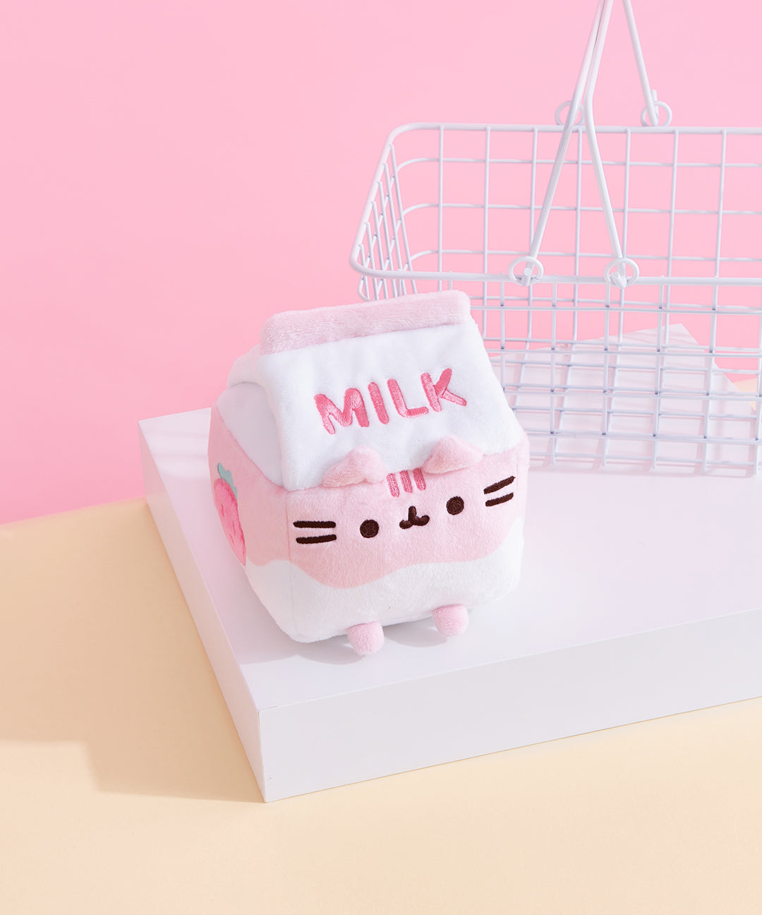 Against a pink background and yellow surface, Pusheen Strawberry Milk Sips Plush is seated on a white pedestal with a small white wire basket in the background. The plush is in the shape of a milk carton, with the word ‘Milk’ embroidered in pink stitching on the front lid of the plush, and a light pink and white wave throughout the body. Pusheen’s face is embroidered in black on the plush, with plush ears at the rim of the carton lid and pink plush feet at the bottom. 
