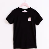 Front view of the Strawberry Milk Sips Tee. The black T-shirt is hanging on a light pink hanger in front of a white background. 
