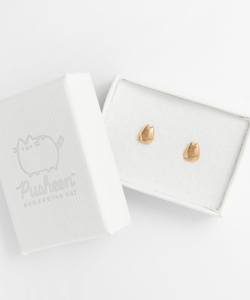The Gold Pusheen Charm Stud earrings in a rectangular white jewelry box, the lid resting on top of the bottom left corner. The gold earrings are the only bit of color in the whole piece, and the earrings reflect the same way the silver earrings do.