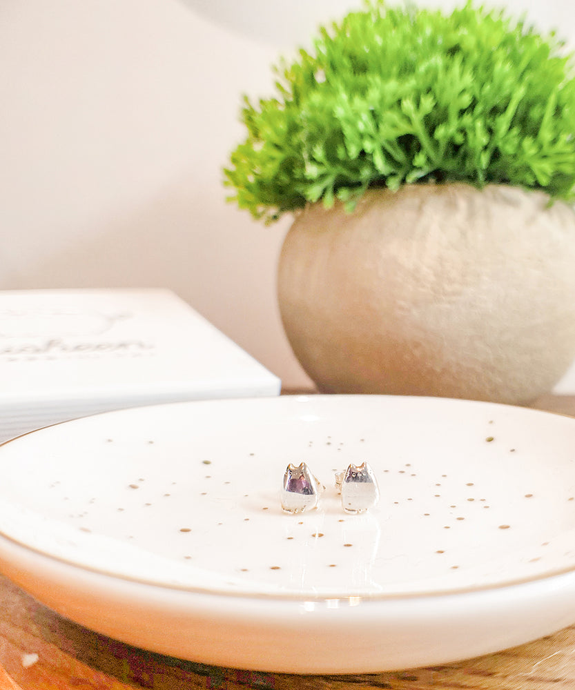 The Silver Pusheen Stud Earrings displayed on a speckled spa dish, on top of a wooden table. Behind the dish is the jewelry box with the Pusheen Logo, as well as a round potted plant. The earrings reflect the dish underneath them.