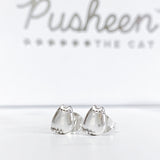 Close up of the Silver Pusheen Stud Earrings, with a hazy view of the Pusheen the Cat logo on the jewelry box lid behind them, appearing larger than actual size. Pusheen’s dot eyes, mouth, and whiskers are present, and faint lines depicting Pusheen’s forehead stripes are partially visible in between her ears. Pusheen’s feet and tail at he bottom are extra dots of silver, and Pusheen’s tail have some faint stripes. The earrings use a pushback backer.