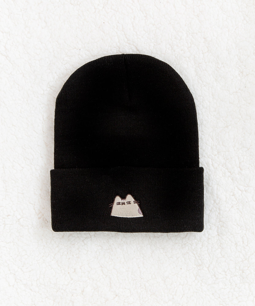Top view of the Suspicious Pusheen Knit Hat. Black beanie lays on top of a fluffy white carpet. On the front center of the folded edge is an embroidered  Pusheen the Cat looking hesitantly to the side. 