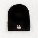 Top view of the Suspicious Pusheen Knit Hat. Black beanie lays on top of a fluffy white carpet. On the front center of the folded edge is an embroidered  Pusheen the Cat looking hesitantly to the side. 