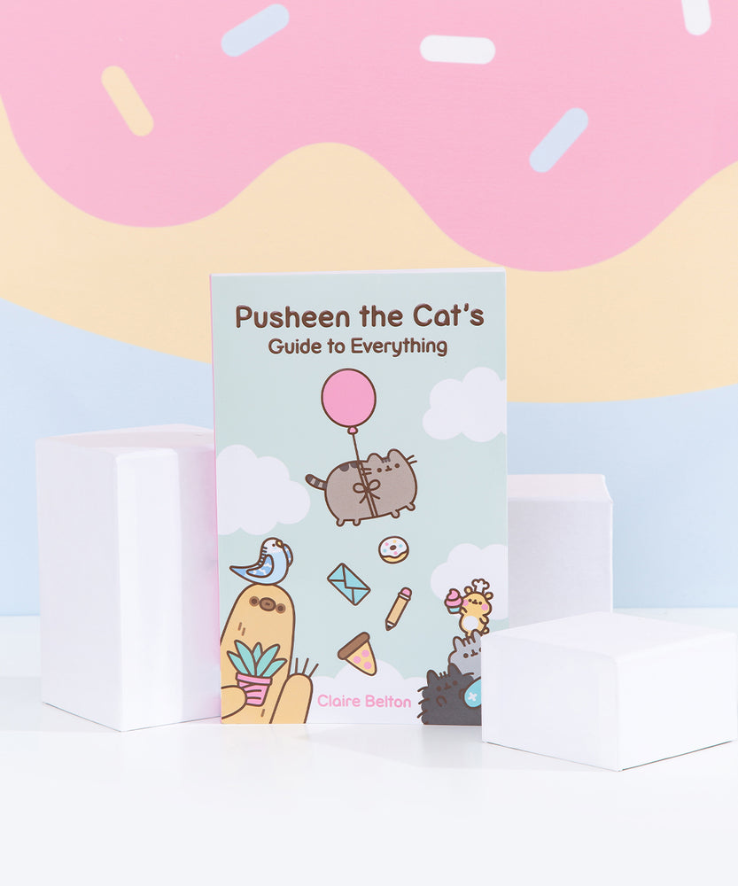 Front view of Pusheen the Cat's Guide to Everything. Little blue book features Pusheen, Bo the Parakeet, Sloth, Cheek the Hamster, Little Sister Stormy, and Little Brother Pip characters on the cover artwork. The propped up book is surrounded by three white cubes while the pink, yellow, and light blue background emulates an ice cream cone drip with sprinkles.