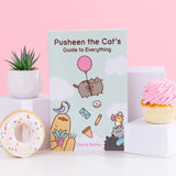 Pusheen the Cat's Guide to Everything paperback book. Blue book features Pusheen, Bo the Parakeet, Sloth, Cheek the Hamster, Little Sister Stormy, and Little Brother Pip characters on the cover artwork. The propped up book is surrounded by three white cubes with a succulent, white glazed donut, and pink cupcake.