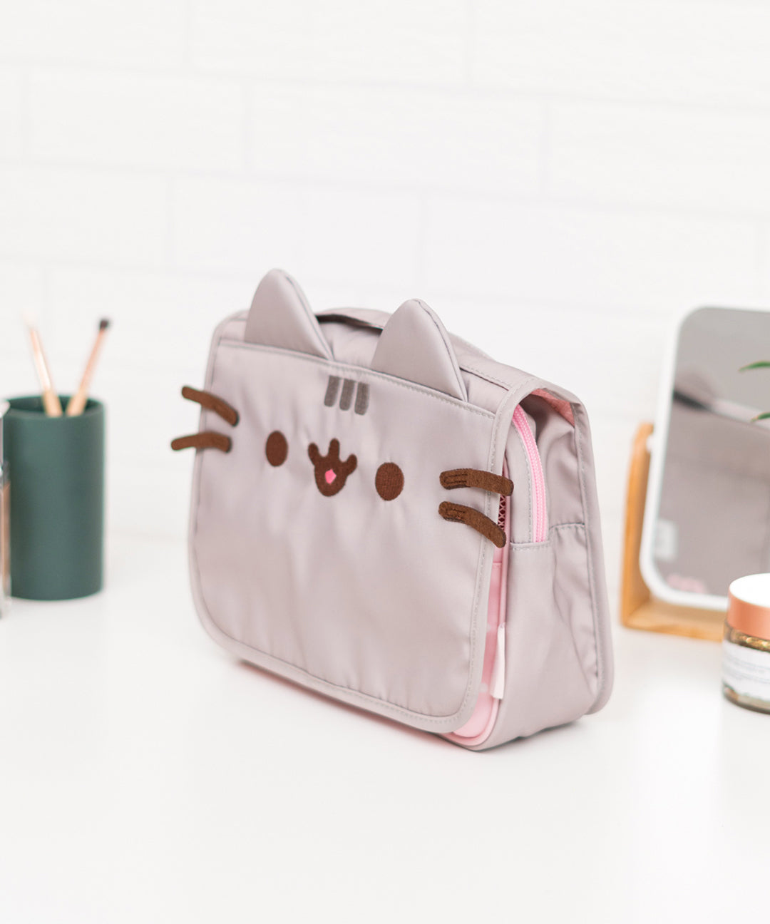 Quarter front view of Pusheen Toiletry Bag. The grey polyester bag features Pusheen the Cat with accompanying grey ears, stripes, and brown facial embroidery. The bag has a top handle for carrying.  