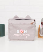 Back view of the grey toiletry bag. The back has a storage slot. Below the storage slot is a saying “Bon Meowage” in white and pink print. The “O” in Bon is a two-toned pink glazed donut.  