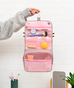 Model holds the toiletry bag open. The bag features a top hook for hanging. The interior of the tri-fold bag is light pink pola dot print and has zip-close and mesh storage compartments. The bag closes via magnets at the interior bottom of the bag. 