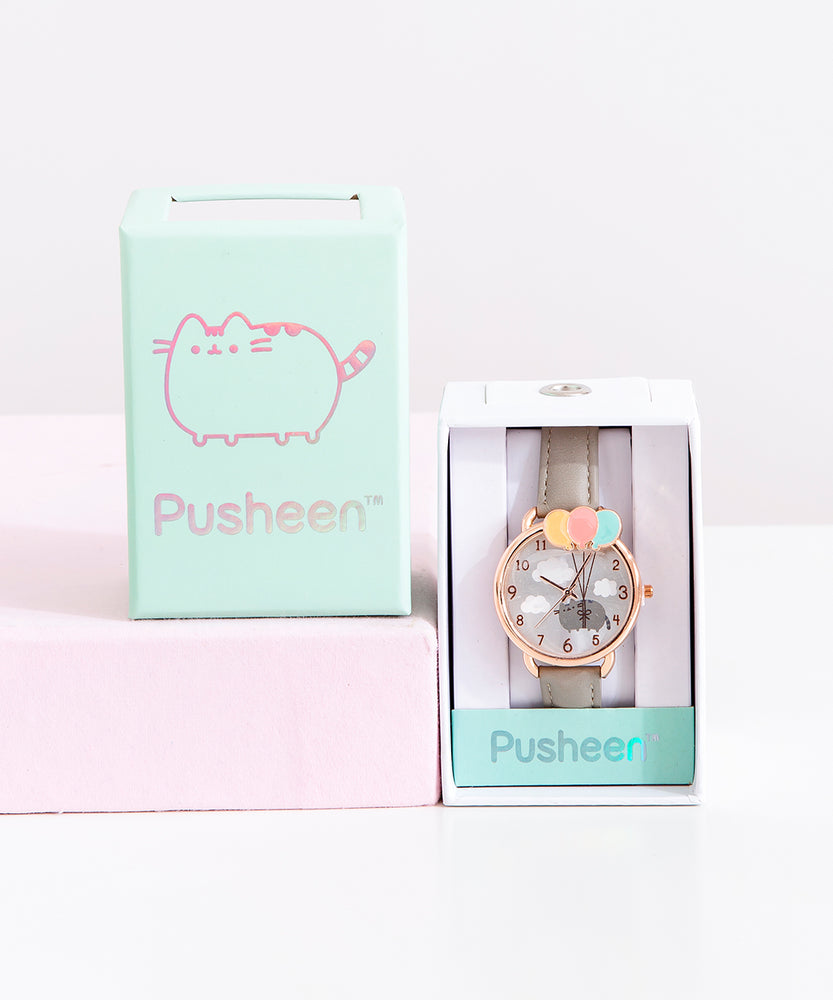 The Pusheen Watch in it’s packaging, with the lid besides it on top of a pink pillar. The watch comes in a white rectangular box, with a mint strip of paper the name ‘Pusheen’ printed in reflective lettering. The lid of the box is the same mint, and features an outline of Pusheen along with the name Pusheen, all in reflective ink.