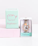 The Pusheen Watch in it’s packaging, with the lid besides it on top of a pink pillar. The watch comes in a white rectangular box, with a mint strip of paper the name ‘Pusheen’ printed in reflective lettering. The lid of the box is the same mint, and features an outline of Pusheen along with the name Pusheen, all in reflective ink.