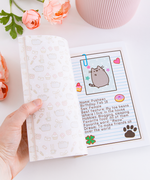 A model’s hand opening the book on top of  white surface, with pink peonies and a pink ribbed pot above the book. The page is turned to Pusheen’s profile, which lists her name, birthday, gender, best feature, hobbies, favorite food, favorite word, and dream. On the opposite page is a repeating pattern of Pusheen among various items like bows, mushrooms, cupcakes, letters, roses and more.