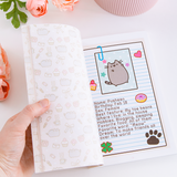 A model’s hand opening the book on top of  white surface, with pink peonies and a pink ribbed pot above the book. The page is turned to Pusheen’s profile, which lists her name, birthday, gender, best feature, hobbies, favorite food, favorite word, and dream. On the opposite page is a repeating pattern of Pusheen among various items like bows, mushrooms, cupcakes, letters, roses and more.