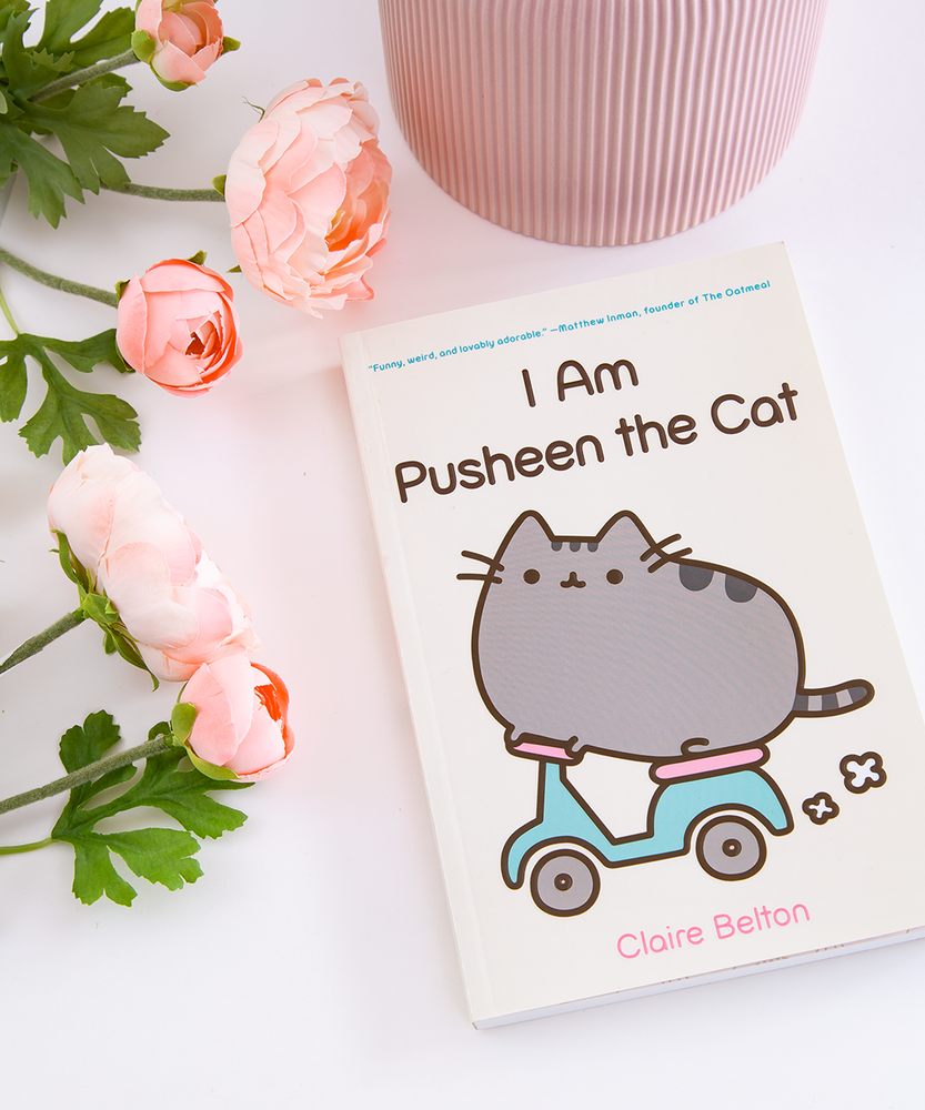 The I Am Pusheen the Cat Book on top of a white surface, with pink peonies and a pink ribbed pot around it.