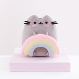 Front view of the Pusheen with Rainbow Plush sitting on top of a pink square pedestal in front of a white background. The ends of the rainbow are rounded out and vary in size, as if the rainbow is three strings of clay placed on top of one another. Pusheen’s bottom two feet peek out from underneath the inner edges of the rainbow.